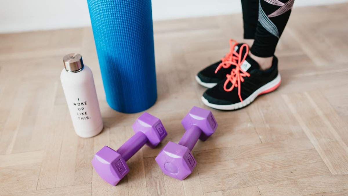 Unrecognizable sportswoman wearing sneakers and black leggings standing with sport mat on wooden floor near dumbbells and water bottle before exercising indoors
